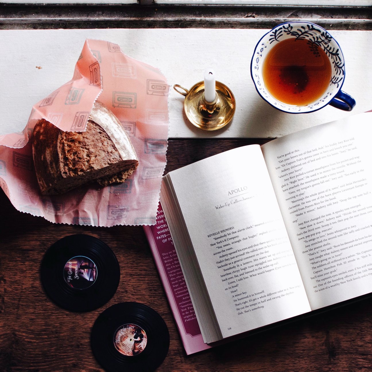 features an open book, a tea cup, beeswax wrap half covering a  half loaf of read. Two vinyl coasters as beside the book