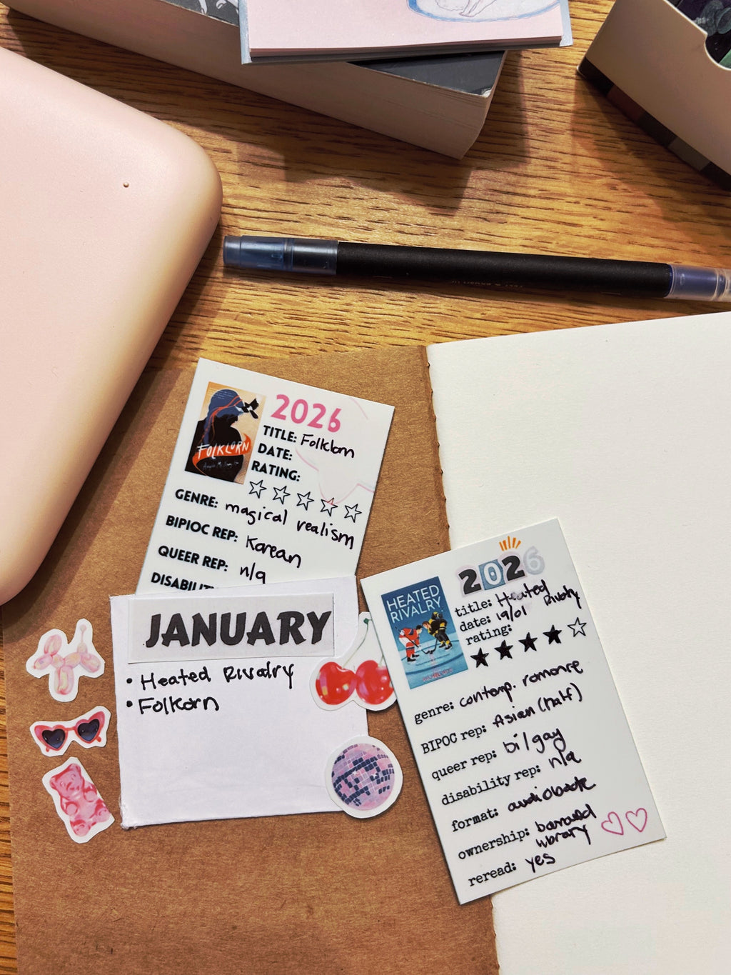 Two reading journal cards on top of a reading journal notebook surrounded by stickers and books.