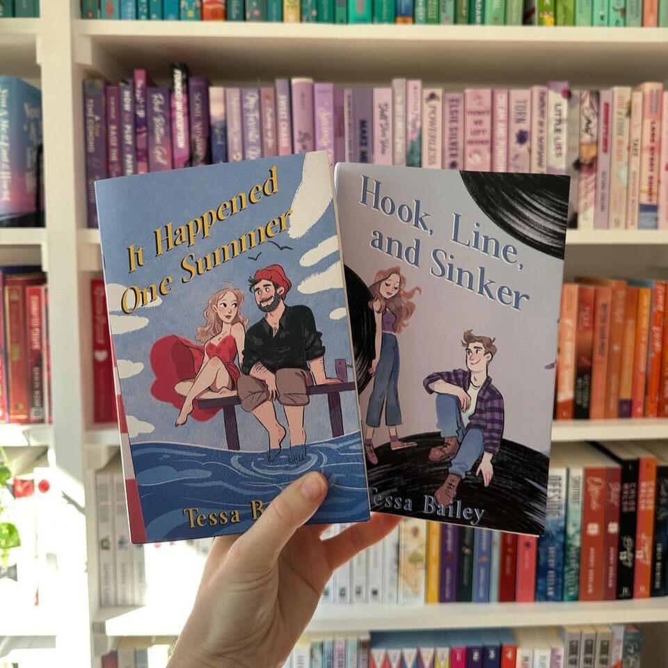 A hand holding two paperbacks of the Bellinger Sisters series by Tessa Bailey, with book titles 'It Happened One Summer' and 'Hook, Line, and Sinker'. The books are in front of a shelf with various colored book spines.