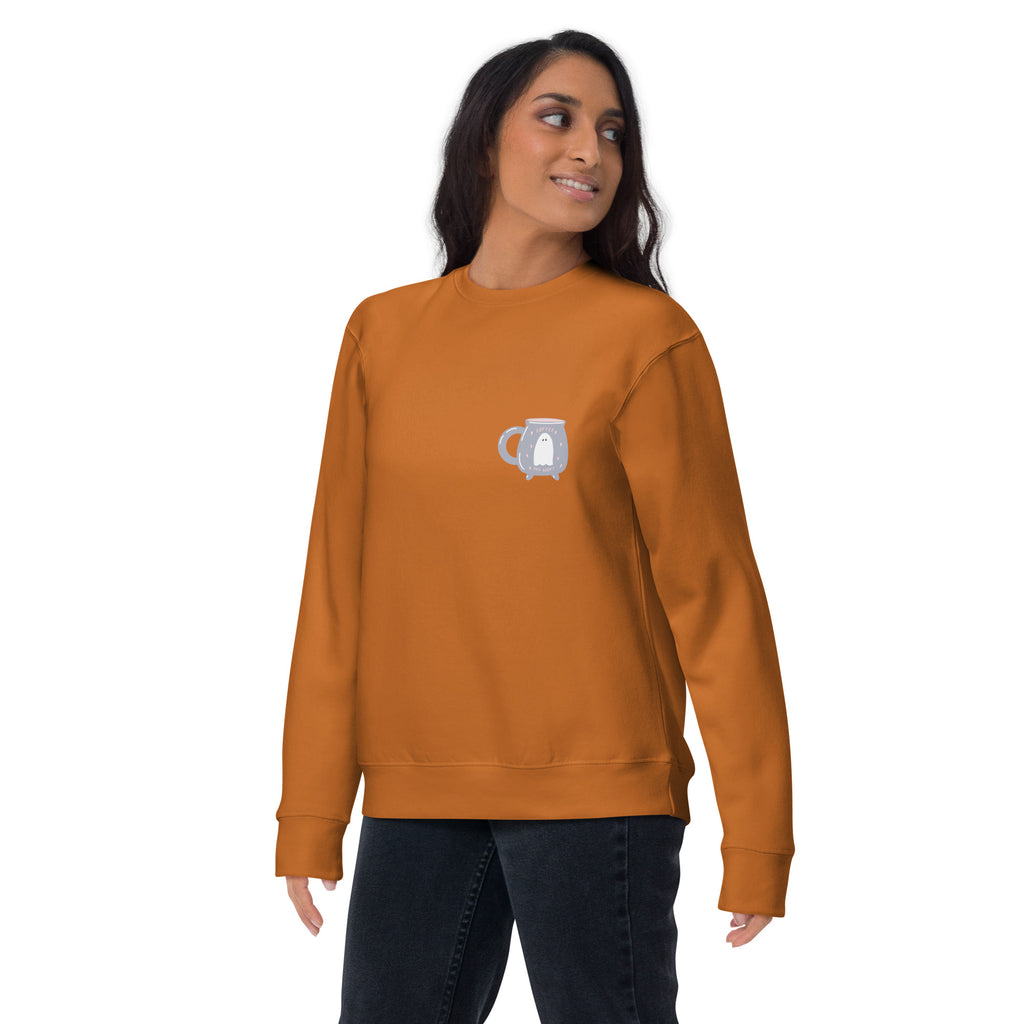 Woman wearing a burnt orange colored sweatshirt with a pocket design featuring a ghost and coffee mug. She is standing against a white back drop.