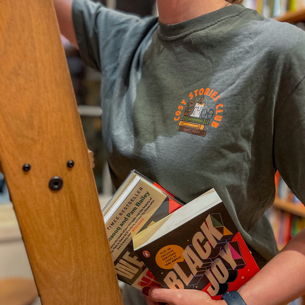 Person holding books with a bookshelf in the background. Woman's t-shirt reads "cosy stories club."