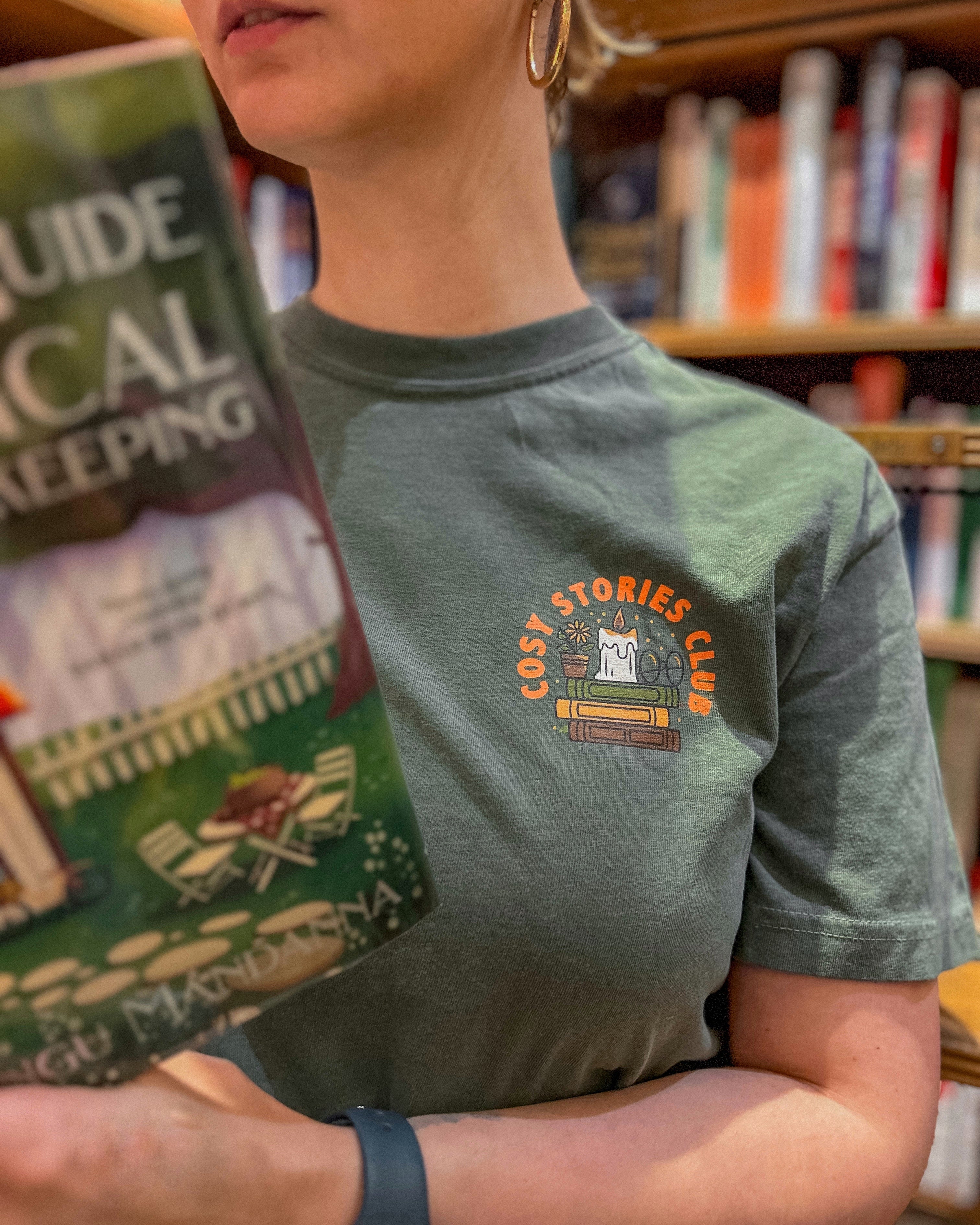 Woman holding a book wearing a green shirt with the pocket design saying "cosy stories club." Standing in front of a bookshelf blurred in the background.