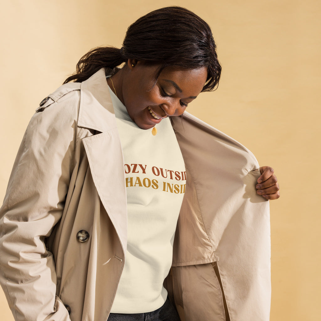 Black woman wearing a cream sweater that reads "cozy outside, chaos inside." She is holding her coat open with her hand and is standing in front of a biege background.