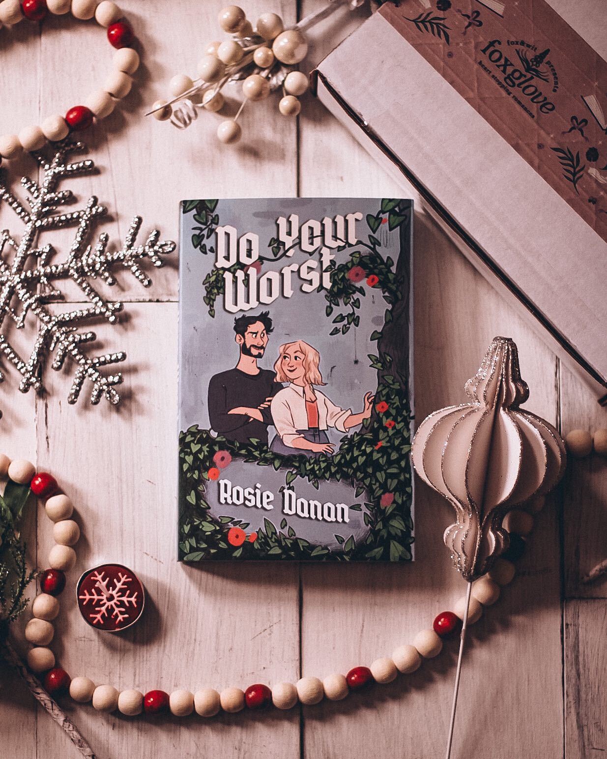 A book titled 'Do Your Worst' by Rose Donahue, featuring an illustration of a man and a woman in a forest setting. The book is placed on a wooden surface with decorative elements around it.