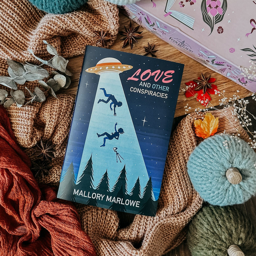 The image displays the book titled "Foxglove: Love and Other Conspiracies" by Mallory Marlowe, presented on a backdrop with a cozy, autumnal vibe, surrounded by leaves, pine cones, and a knitted blanket.