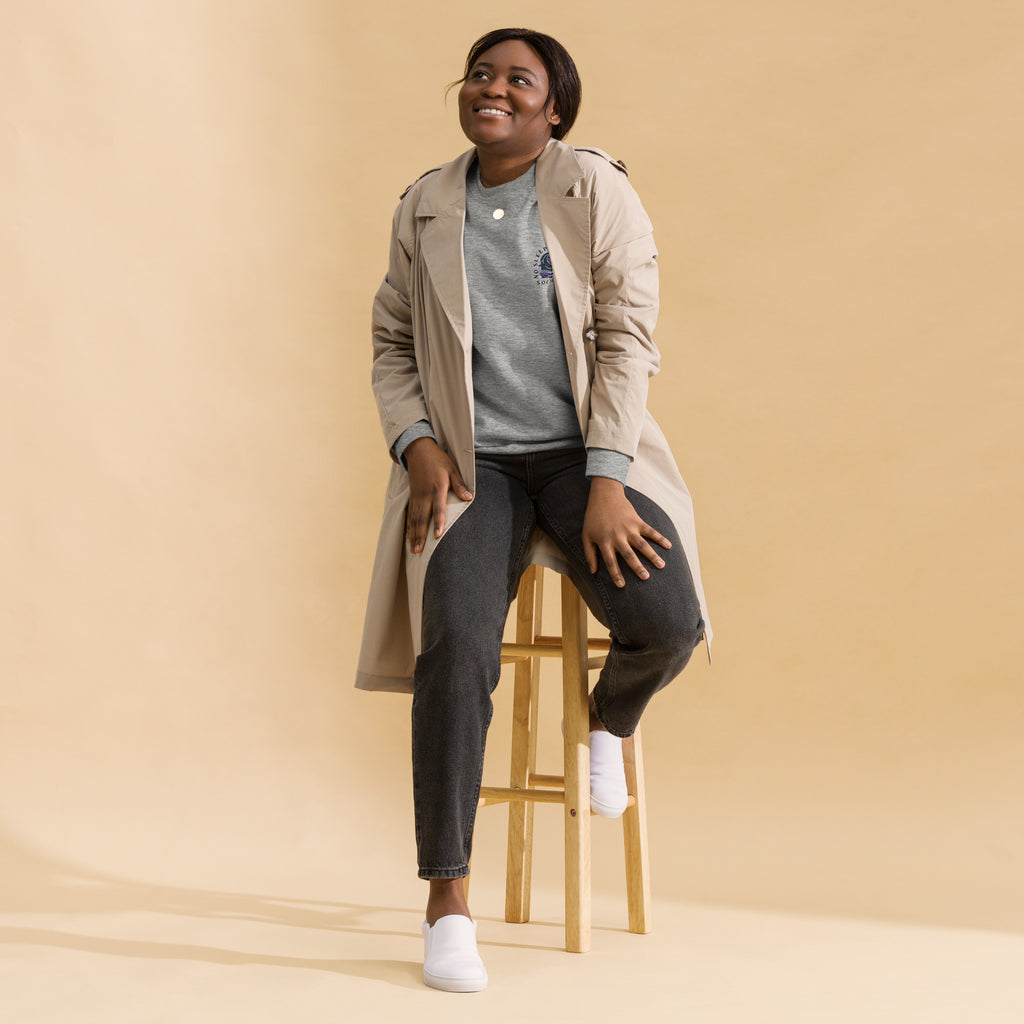 Image of a Black woman wearing a grey sweatshirt  with the design "no sleep reader society" and a cream trench coat. She is sitting on a chair and wearing black trousers and white sneakers.