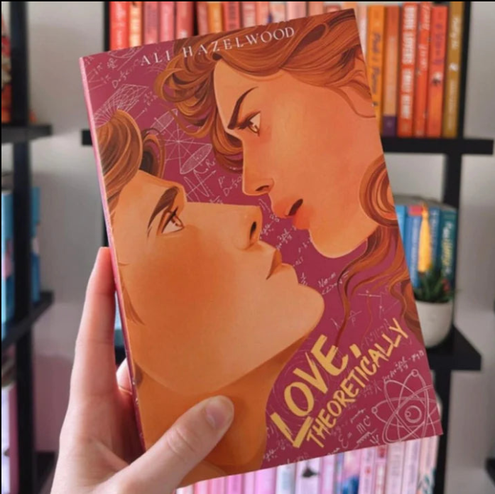 Book titled 'Love Theoretically' held by a hand with a bookshelf in the background