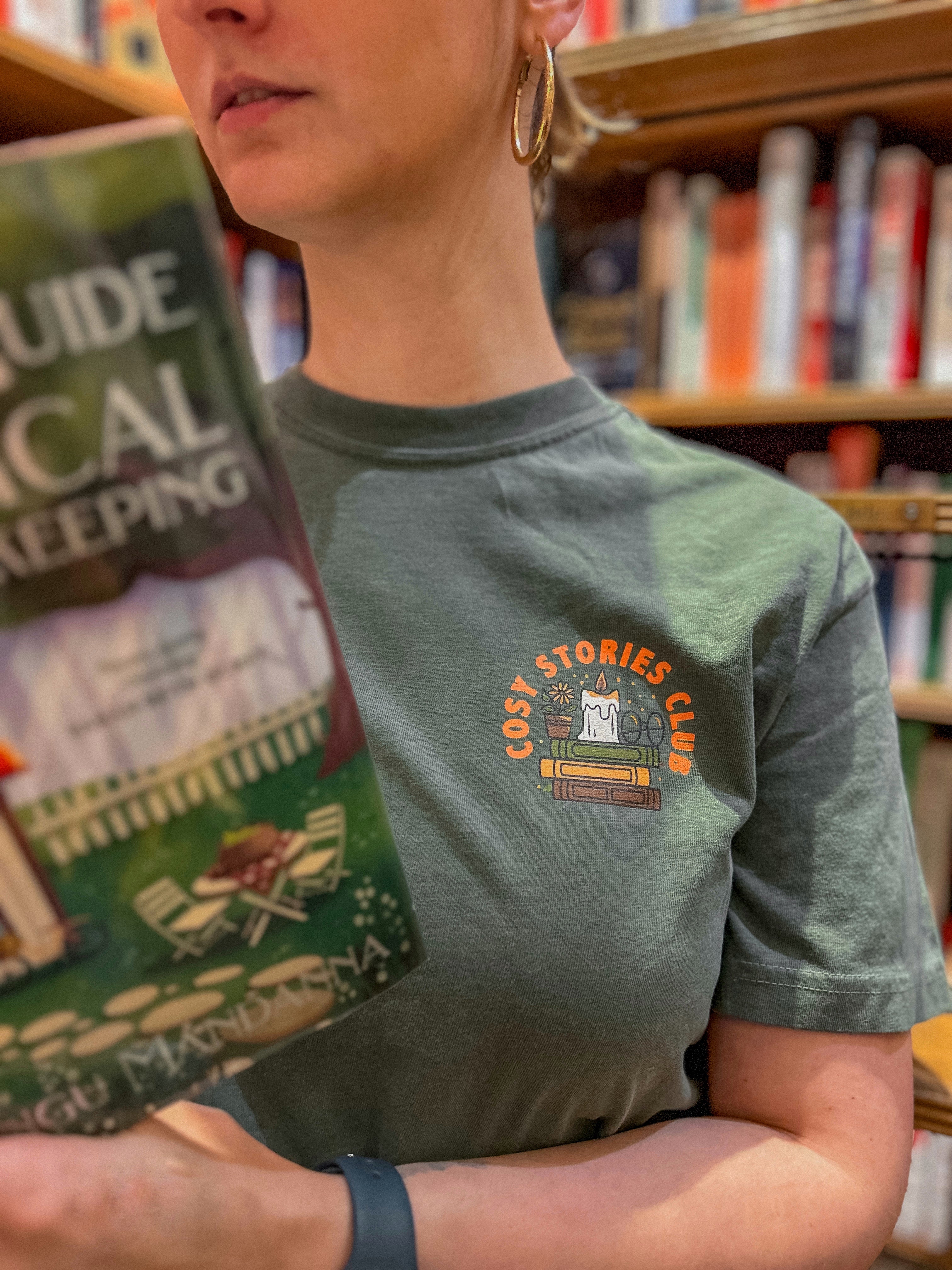 Person wearing a green t-shirt with a 'Cosy Stories Club' design, holding a book in a bookstore.