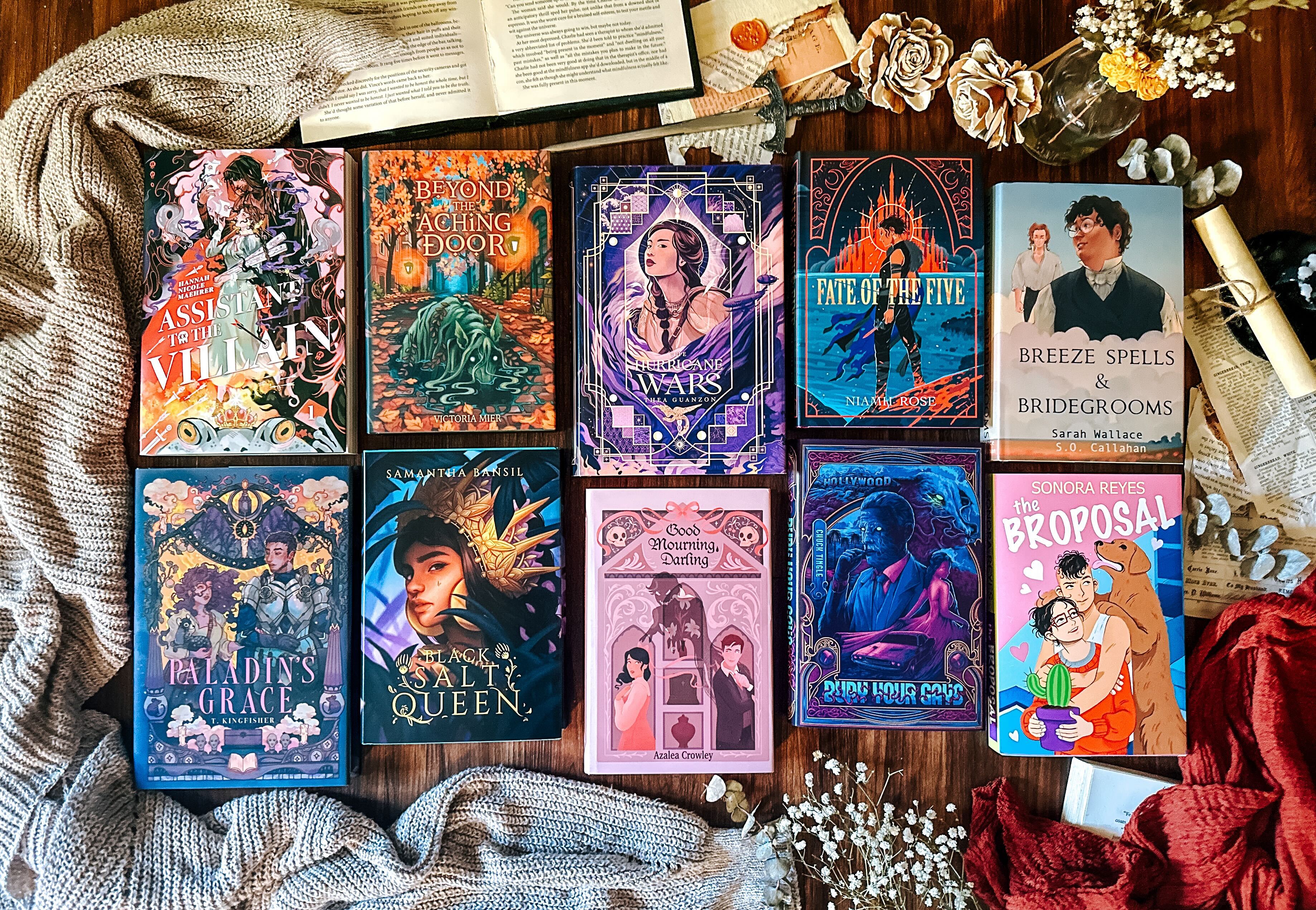 Collection of colorful book covers on a wooden surface with decorative elements. Book titled Assistant to the Villains, Beyond the Aching Door, the Hurricane Wars, Fate of the Five, Breeze Spells and Bridegroom, Paladin's Grace, Black Salt Queen, Good Mourning Darling, Bury Your Gays, and the Proposal.