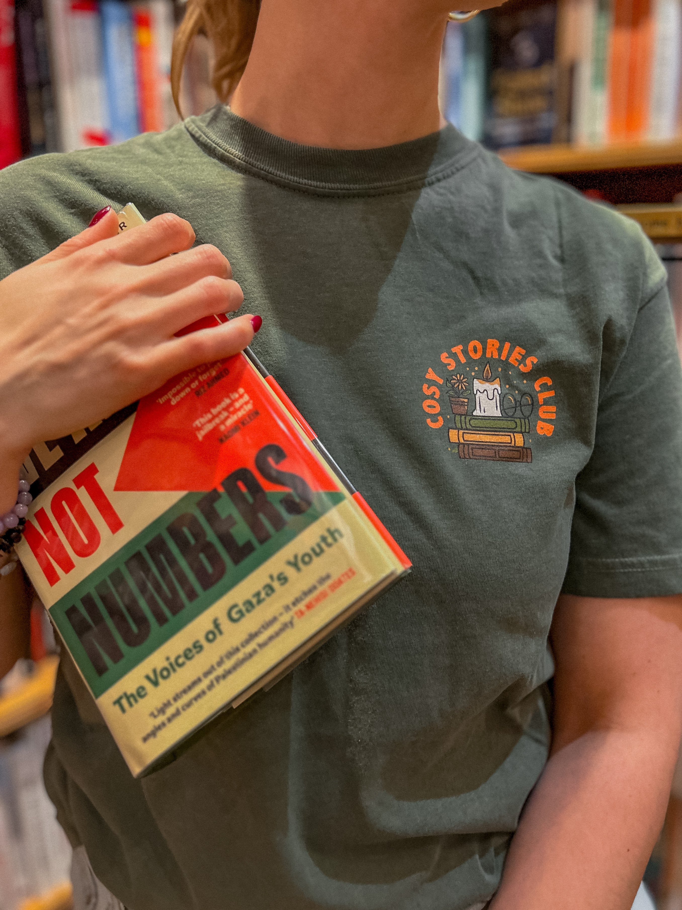 Person wearing a green t-shirt with a design that reads "cosy stories club" and holding book in hand.