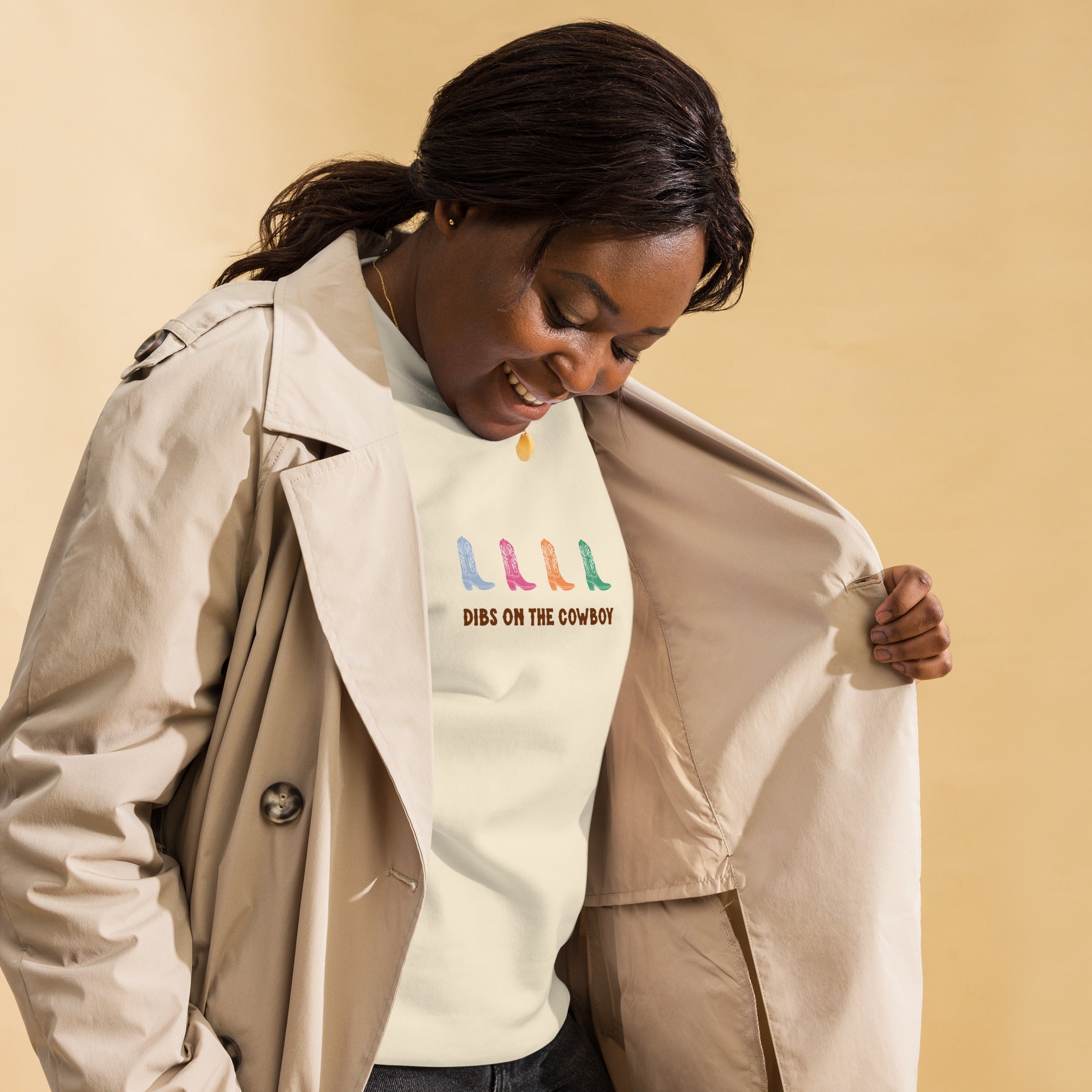 Black woman wearing a beige sweatshirt with four colorful cowboy boots (blue, pink, orange, green) and text that reads "dibs on the cowboy". Shown on a white background