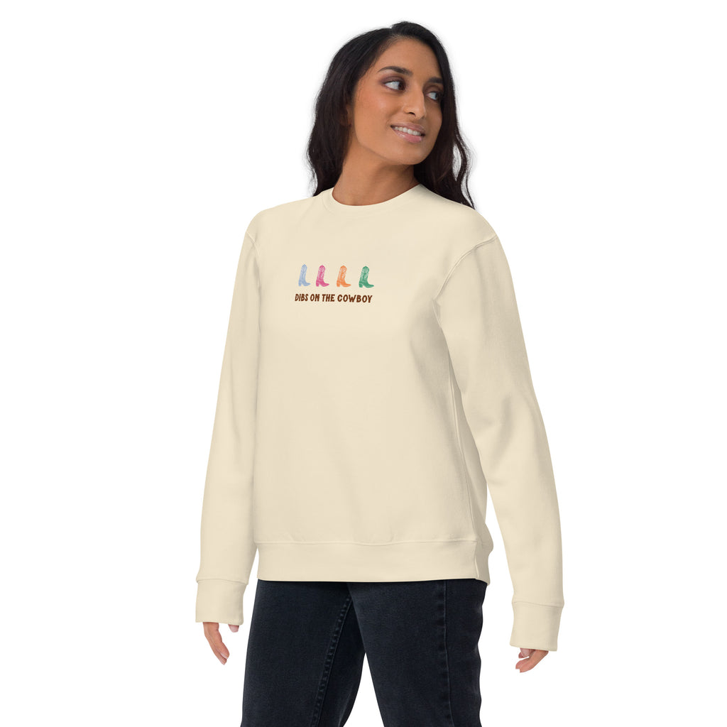 Woman wearing a beige sweatshirt with four colorful cowboy boots (blue, pink, orange, green) and text that reads "dibs on the cowboy". Shown on a white background