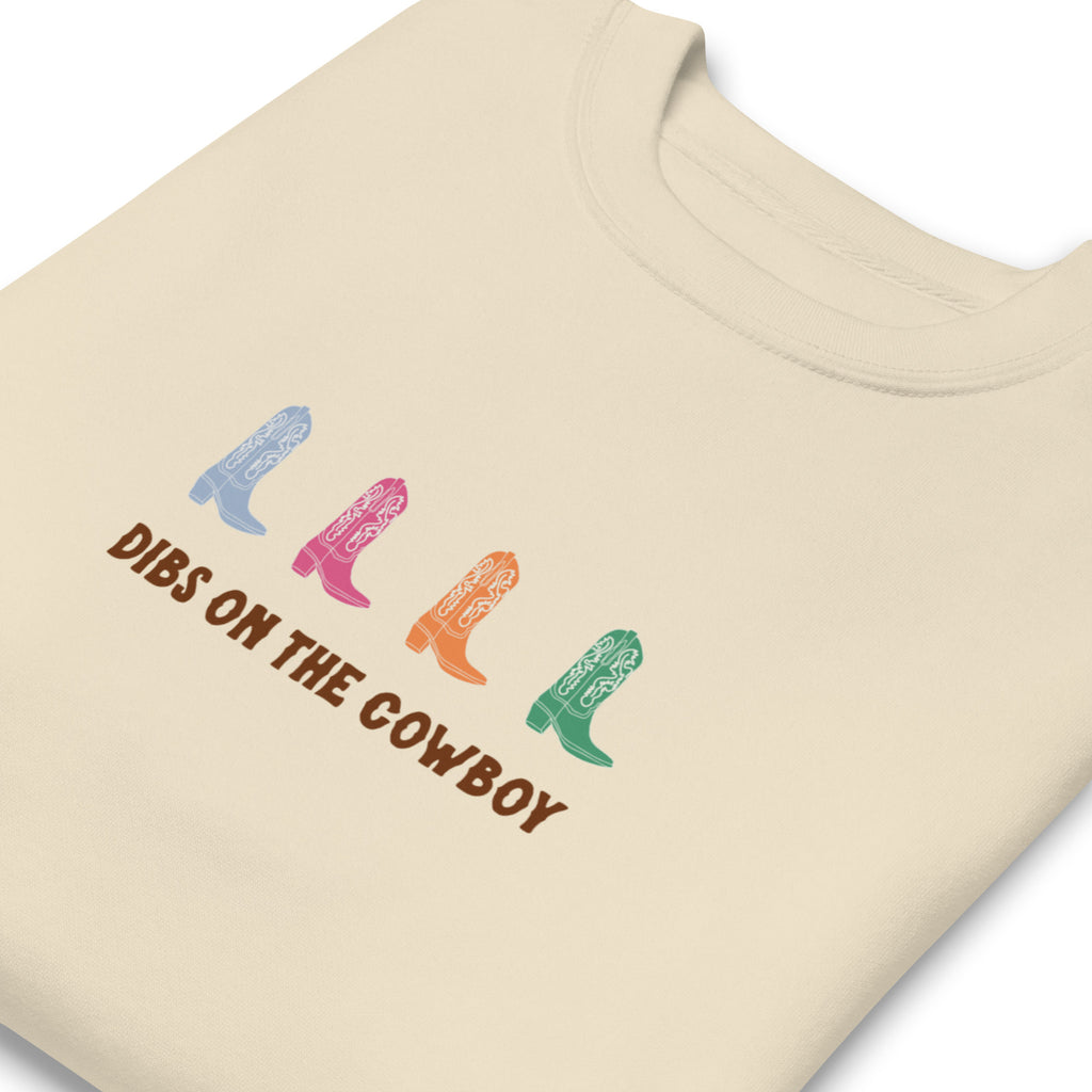 Beige sweatshirt with four colorful cowboy boots (blue, pink, orange, green) and text that reads "dibs on the cowboy". Shown on a white background