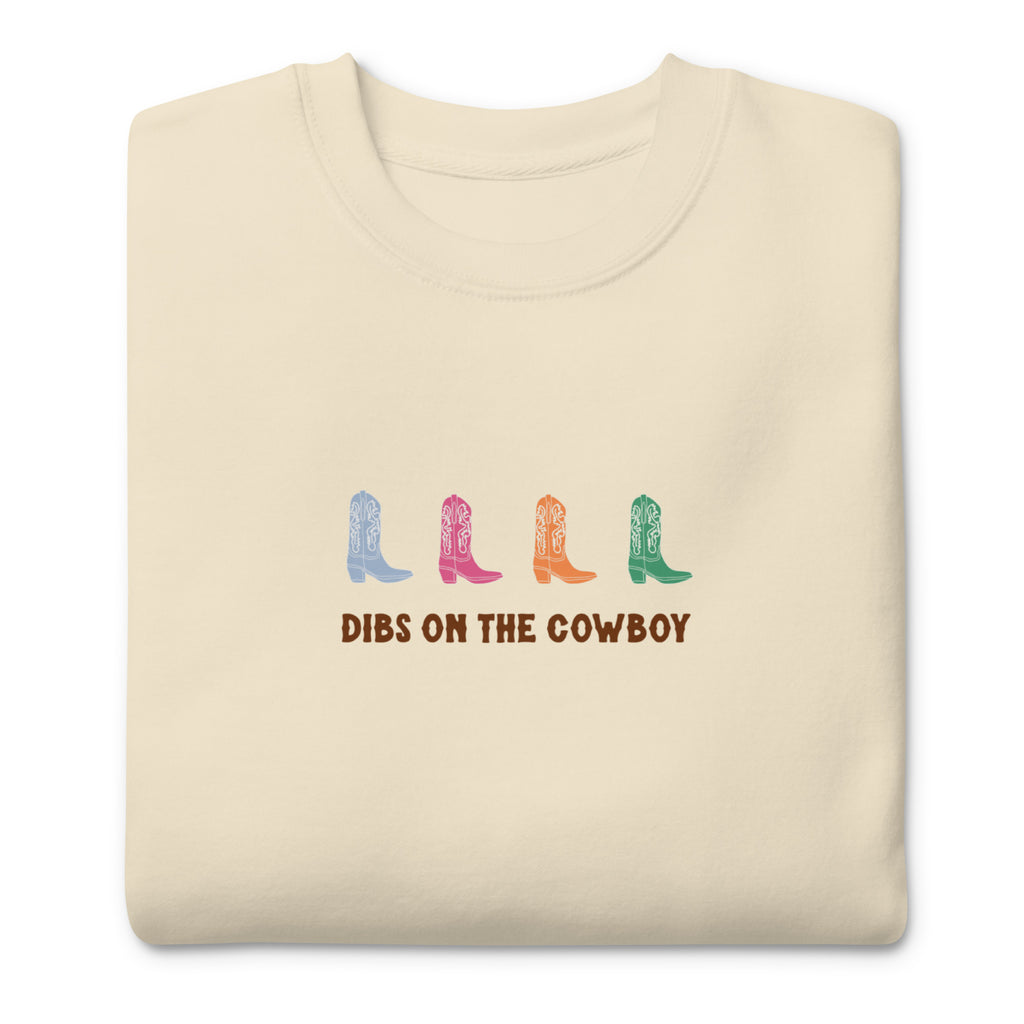 Beige sweatshirt with four colorful cowboy boots (blue, pink, orange, green) and text that reads "dibs on the cowboy". Shown on a white background
