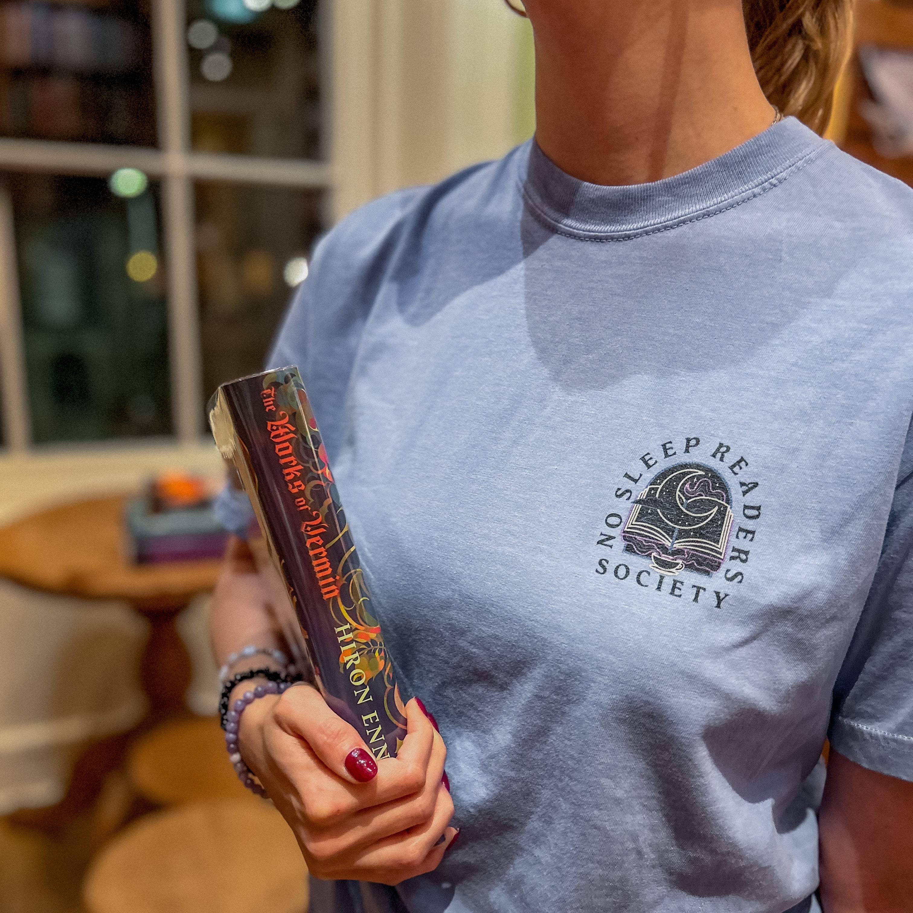 Person wearing a blue t-shirt with a pocket design that reads no sleep reader society and is holding a book in an indoor setting.