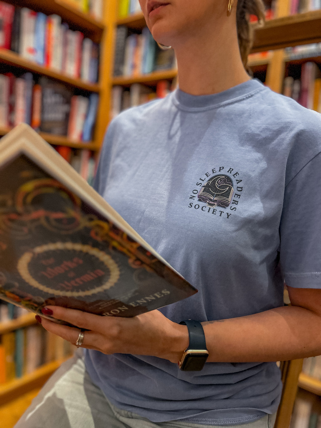 Person wearing a blue t-shirt with a pocket design that reads no sleep reader society and is holding a book in an indoor setting.