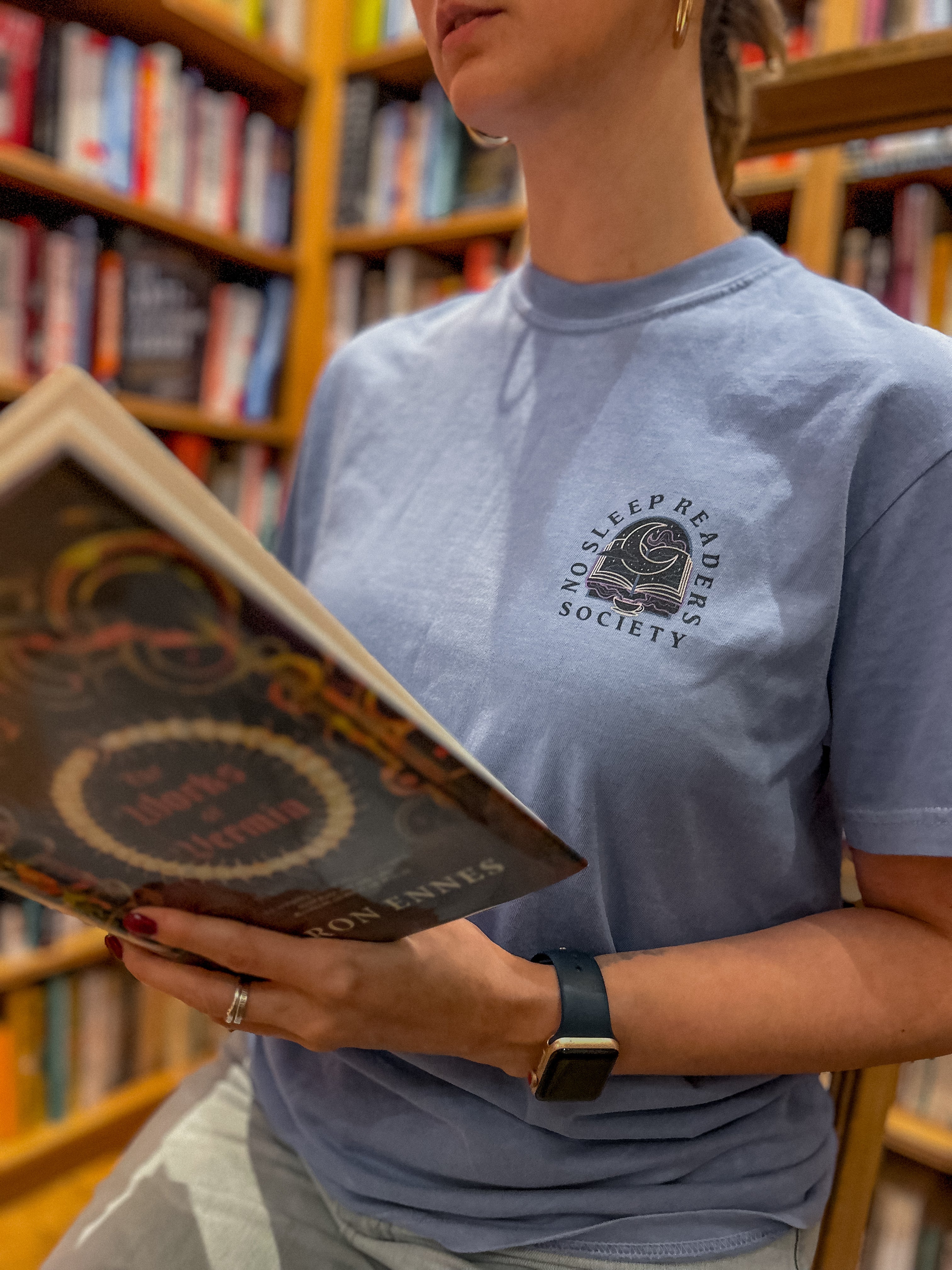 Person wearing a blue t-shirt with a pocket design that reads no sleep reader society and is holding a book in an indoor setting.